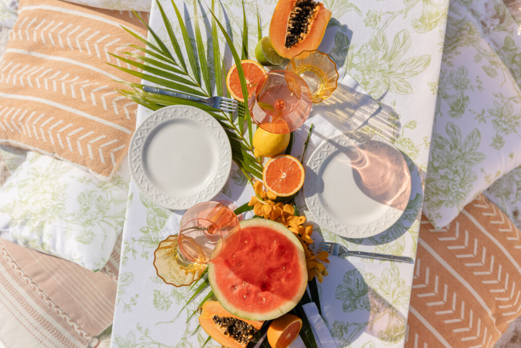 Picnic table on the beach with fruits and pretty pink pillows.